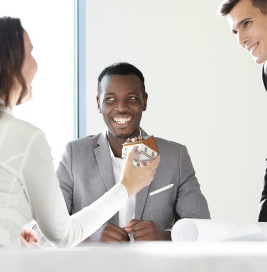 Teamwork and cooperation. Cute female contractor showing her colleagues new real estate project, holding scale model house, presenting design of future building while men looking at her with smile